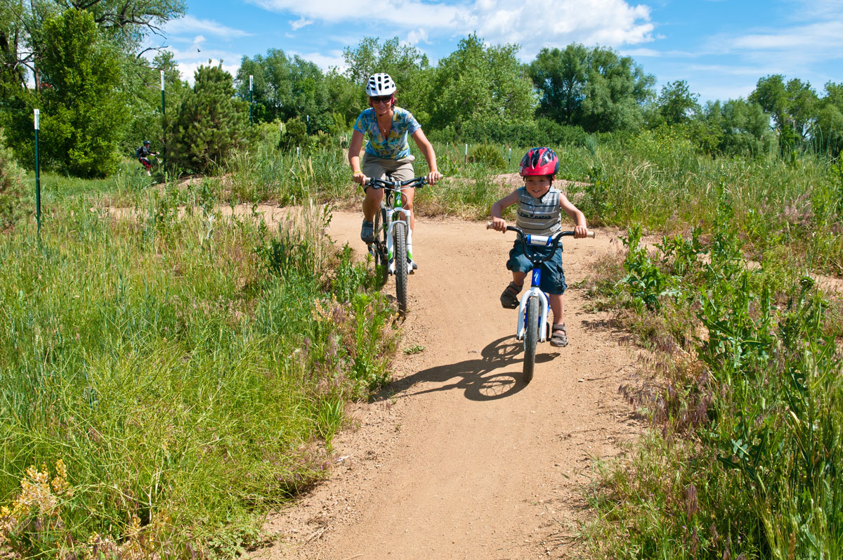 A child and parent ride bikes on a dirt trail surrounded by lush green grass and trees at  Valmont Bike Park in Boulder, Colorado 
