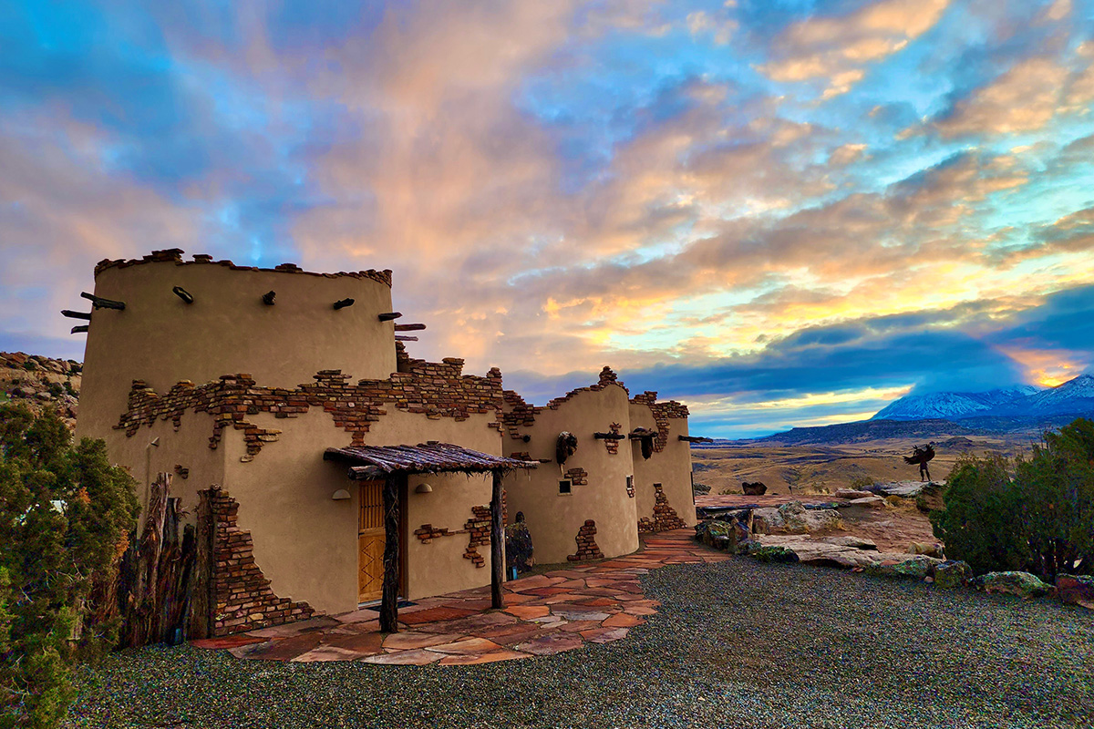 A guesthouse at Canyon of the Ancients Guest Ranch looks like it's made of clay and brick. Beyond the mountains in the background orange and yellow fill the sky with the colors of daybreak.