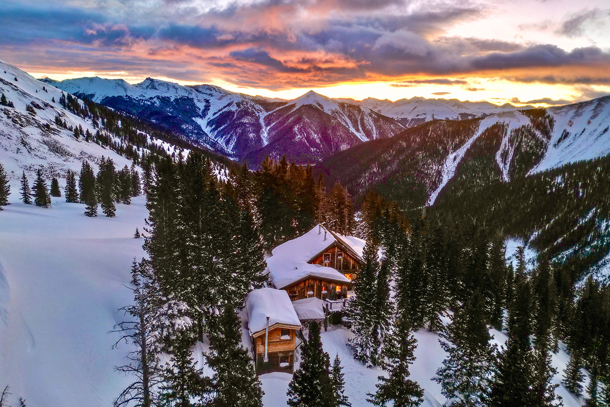 OPUS Hut is blanketed in a thick, fluffy layer of snow as the sun begins to dip below the mountainous Colorado horizon in the distance.