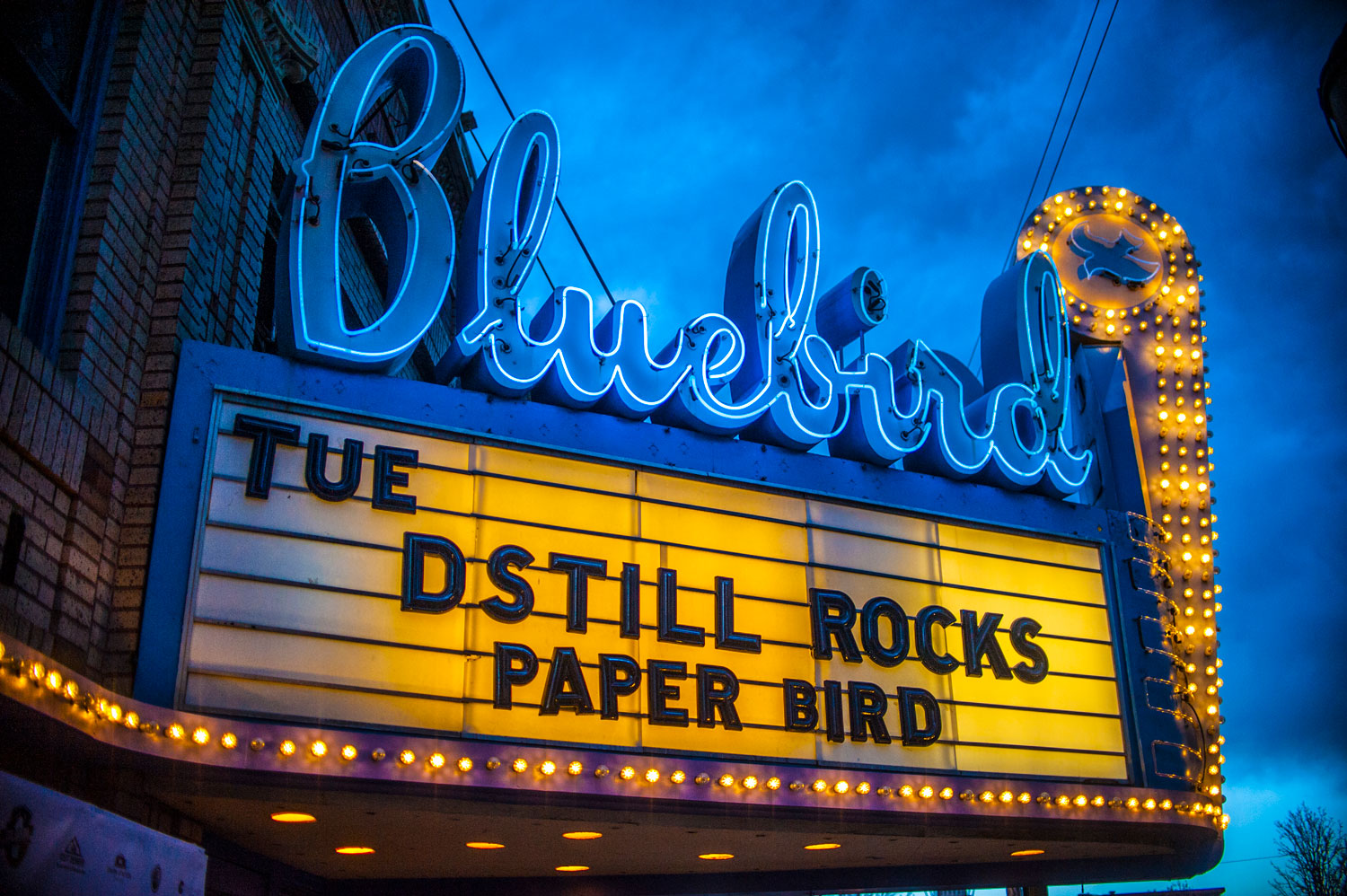 A bright theater marquee lights up against a darkening sky