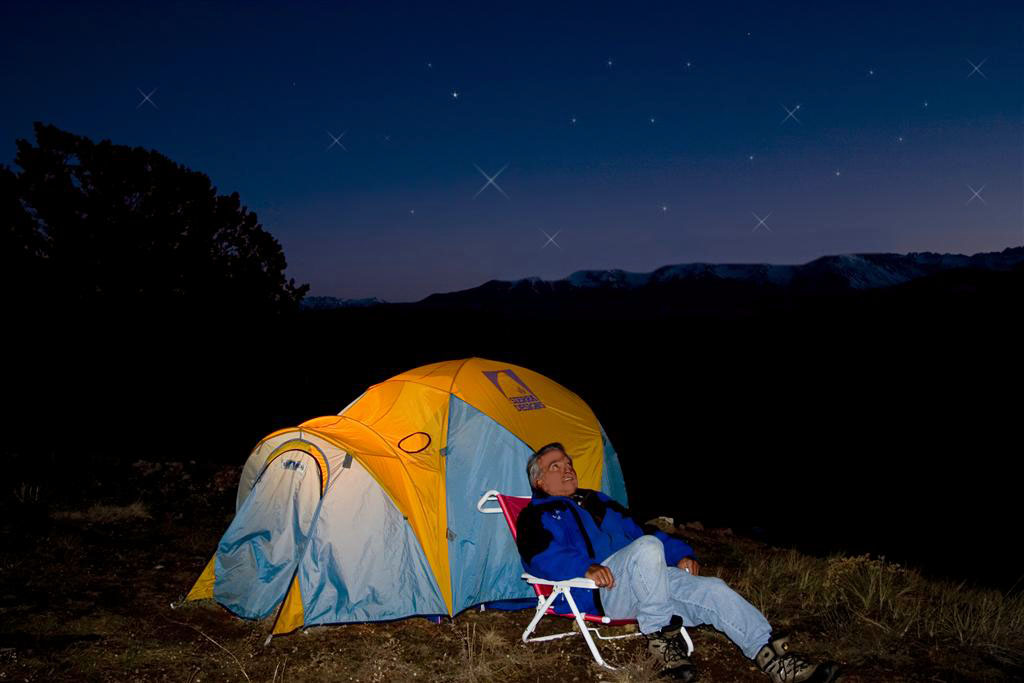 A man in a windbreaker and jeans sits in a camp chair in front of a blue, yellow and white tent. He looks up at the night sky and is surrounded by dark mountains.