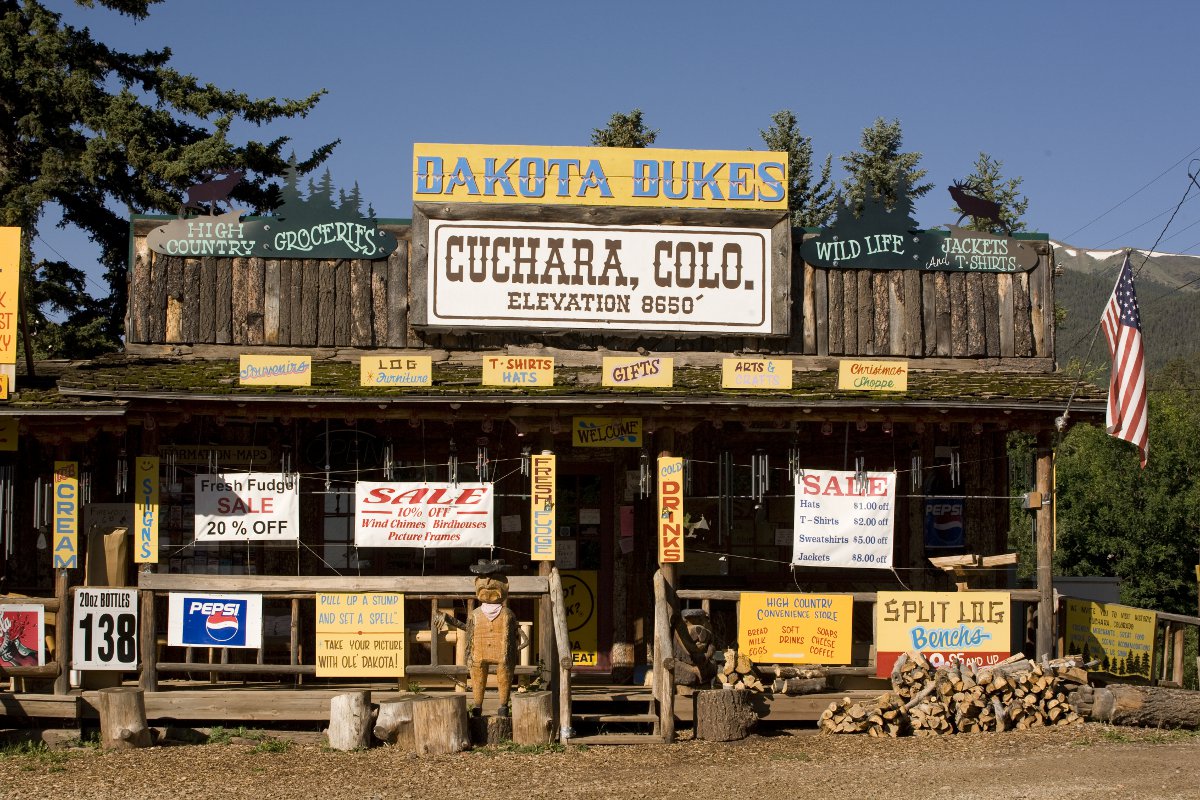 Western storefront with many signs in Cuchara. There's a pile of wood on the right and an American flag hangs off the building.