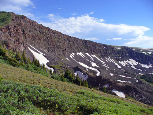 The grey-black ridge of Devil's Causeway is dotted with snow on a blue day with white clouds and there are green grasses on the left.