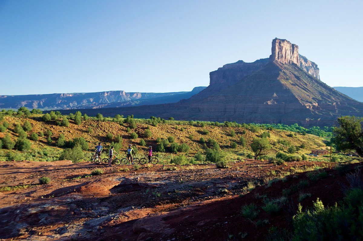 Four mountain bikers look on at a rocky geologic formation that has a butte. There are brushy green bushes, red rocky dirt and the sky is blue. 