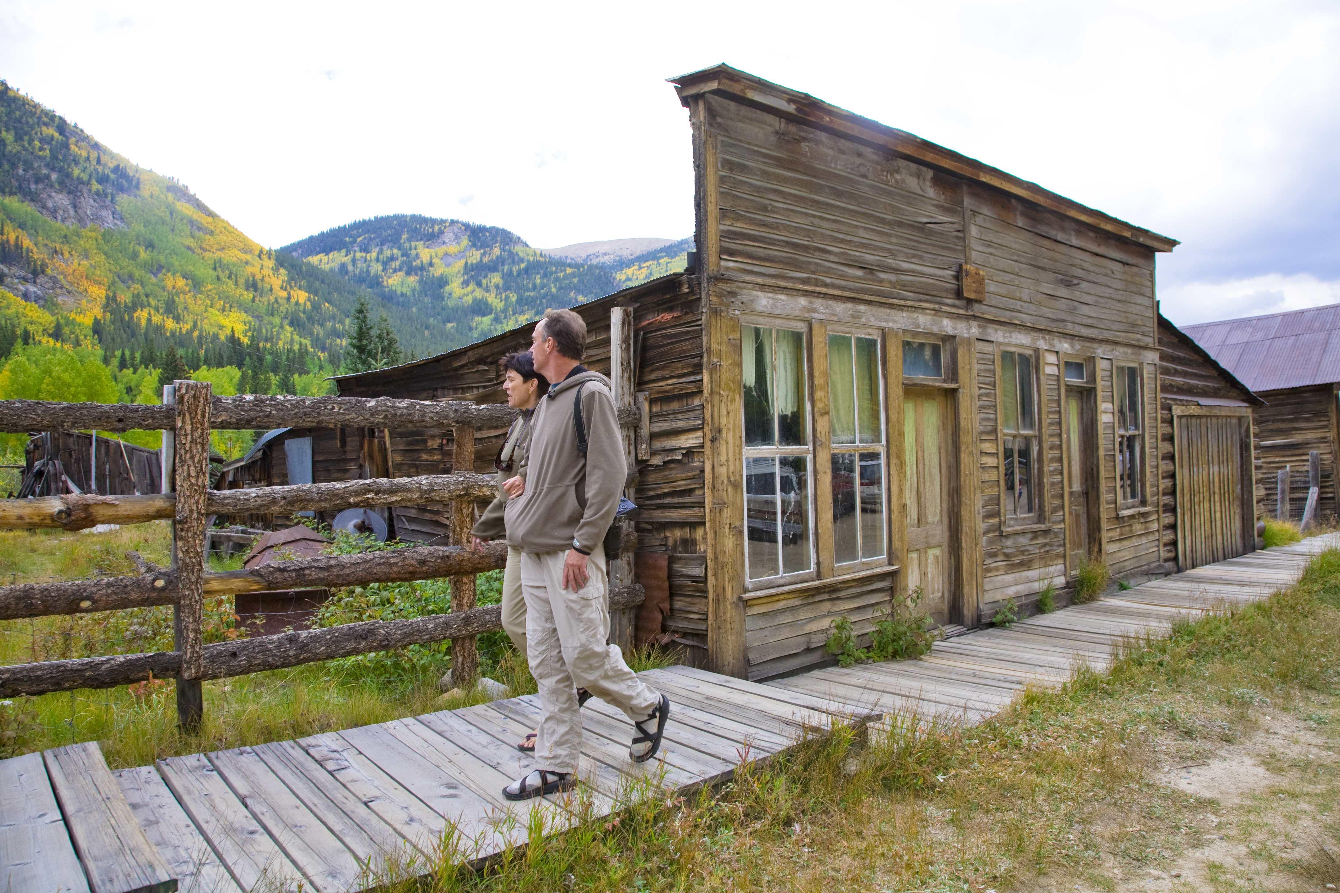 A couple walks on a boardwalk through green grasses passed a wooden storefront at St. Elmo ghost town. In the background, mountains covered in green and golden trees.
