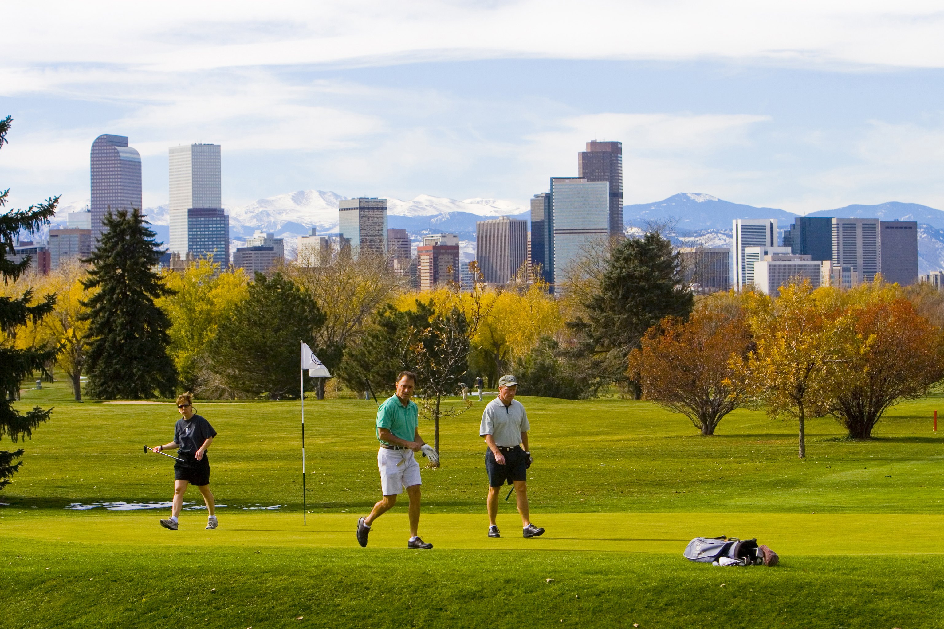 Three men in summer gear walk the green fairways of the golf course. From this view, we can see the many skycrapers of downtown Denver and snowcapped peaks farther west.