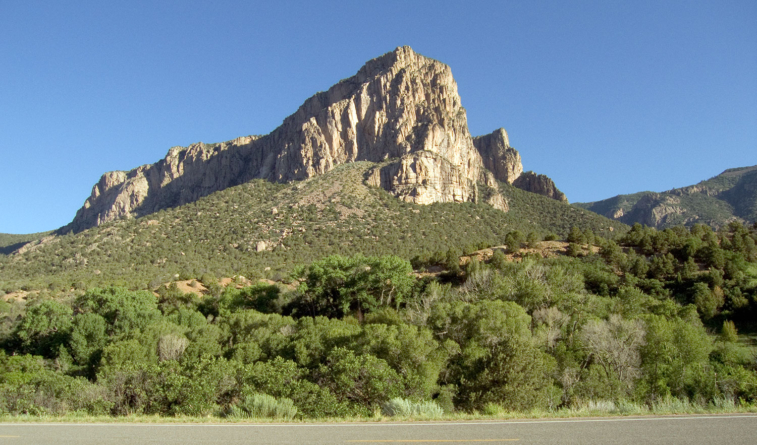 The blacktop passes buy a rocky outcropping that sits atop scrubby green bushes under a blue sky.