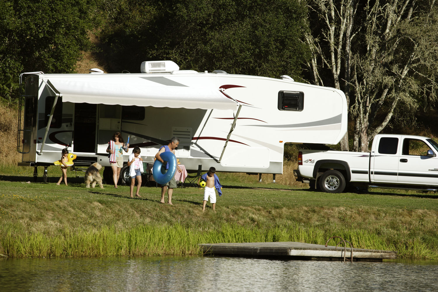 A white RV pulled by a white pickup truck has camp set up, with the awning out, next to a lake. People sit in lawnchairs under the awning.