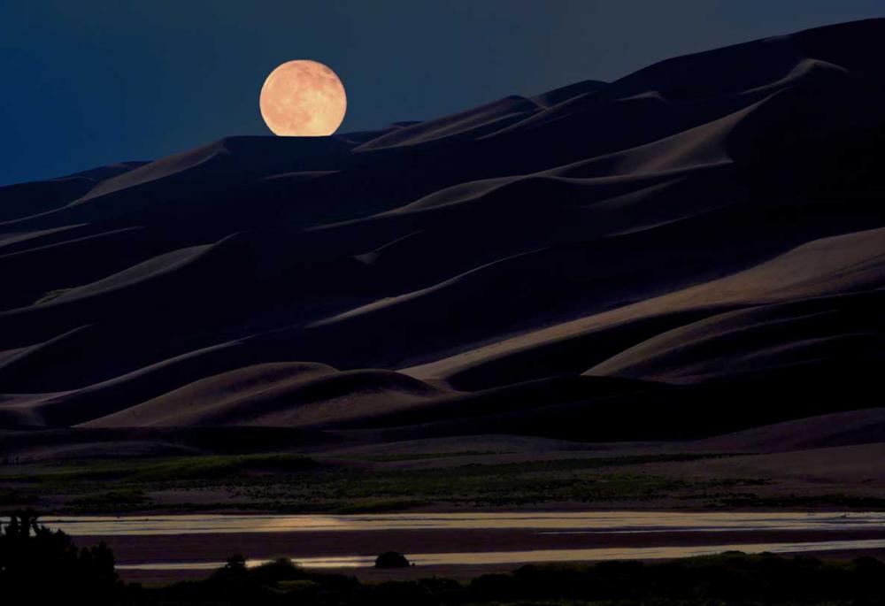 A perfect circle moon sits at night above the Great Sand Dunes.