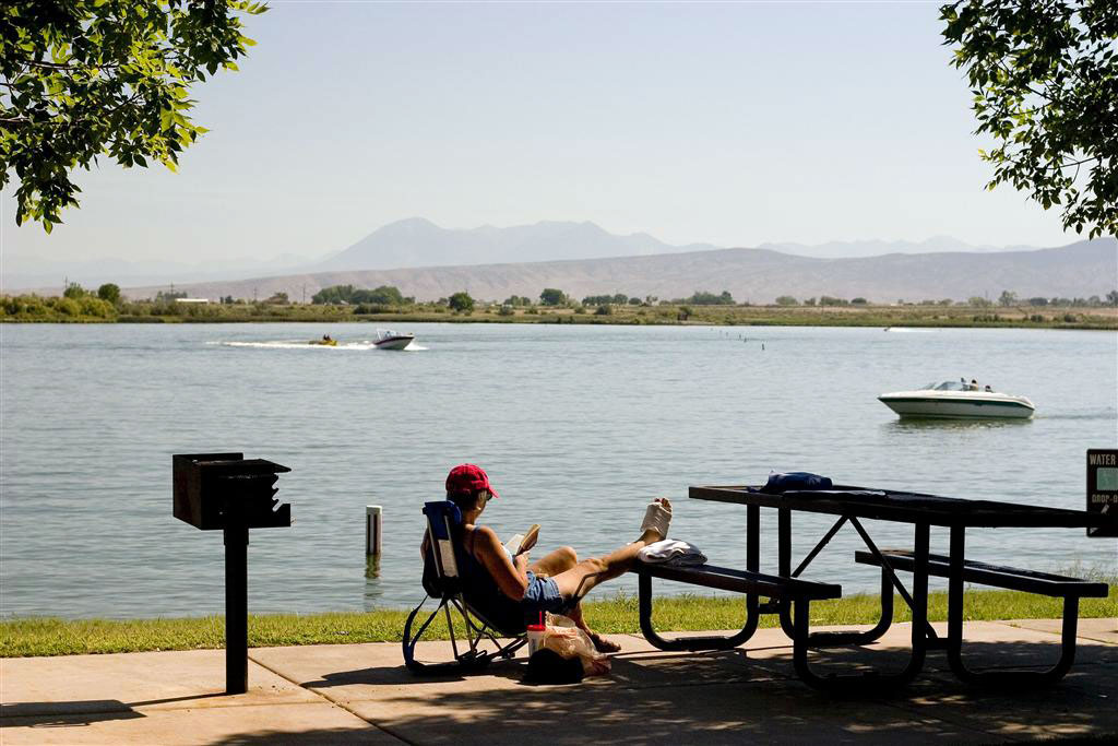 A person sits in a camp chair with their feet propped up on a picnic table on a cement slab with a grill. In the background Sweitzer Lake has two boats with hazy mountains behind them.