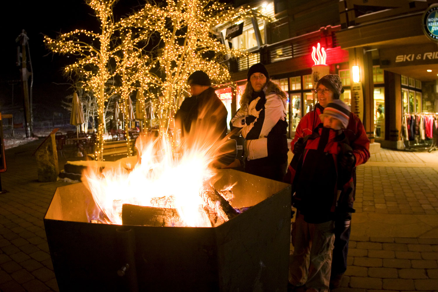 Four people stand in front of a sidewalk fire pit with trees covered in twinkle lights behind them on a Copper Mountain street at night.