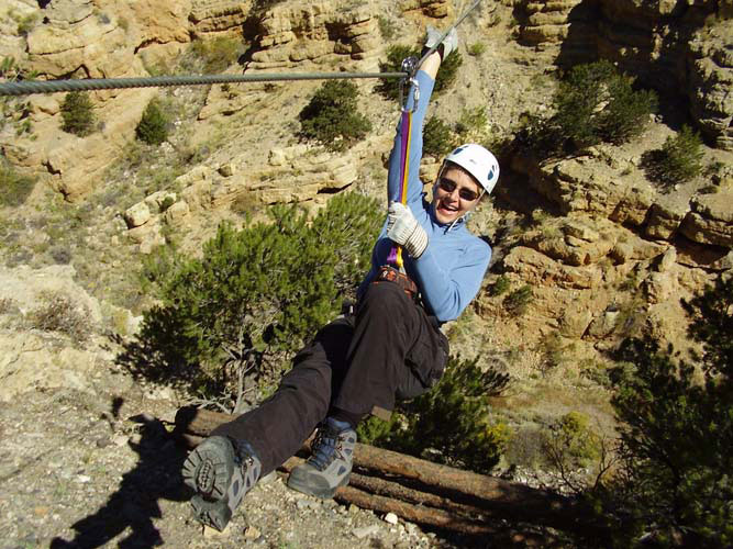 A woman in a helmet tethered to a zipline leans back and prepares to glide across a sagebrush-filled canyon