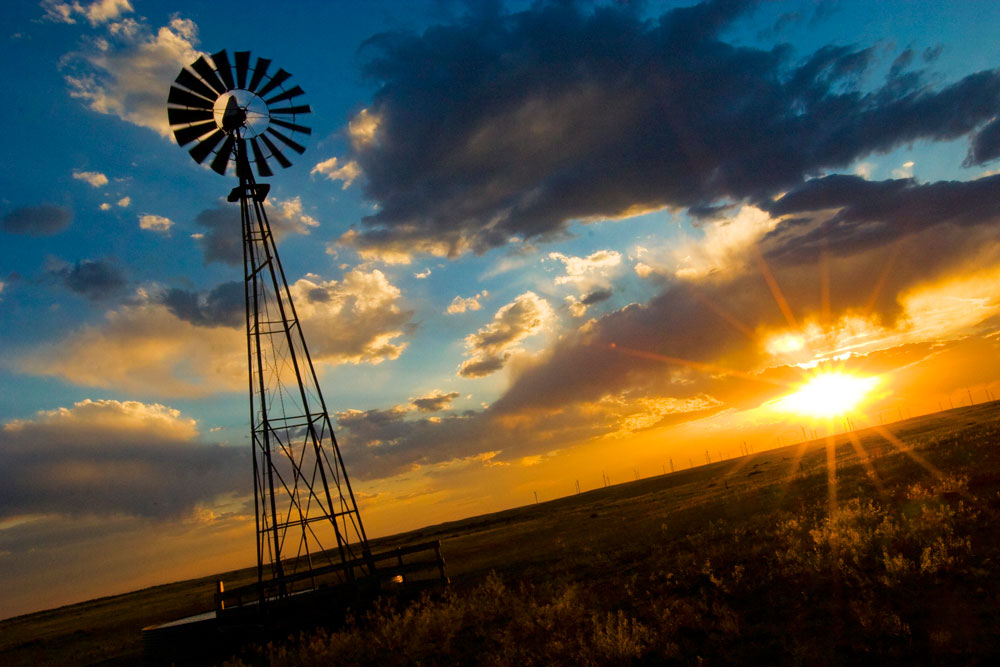 The sun is setting across Colorado's eastern plains with heavy clouds on a blue sky and a lone windmill.