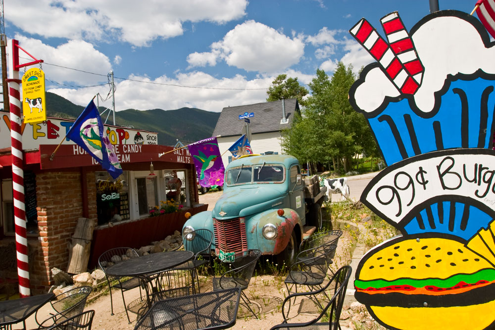 The colorful courtyard of Lewis Sweet Shop sits under a blue sky with white clouds. There's a green with brown spots old truck, a cutout of a milkshake with a burger that reads "99 cent burgers" and metal furniture.