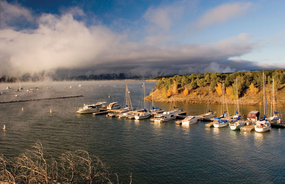A row of sailboats line a doc on a grey lake
