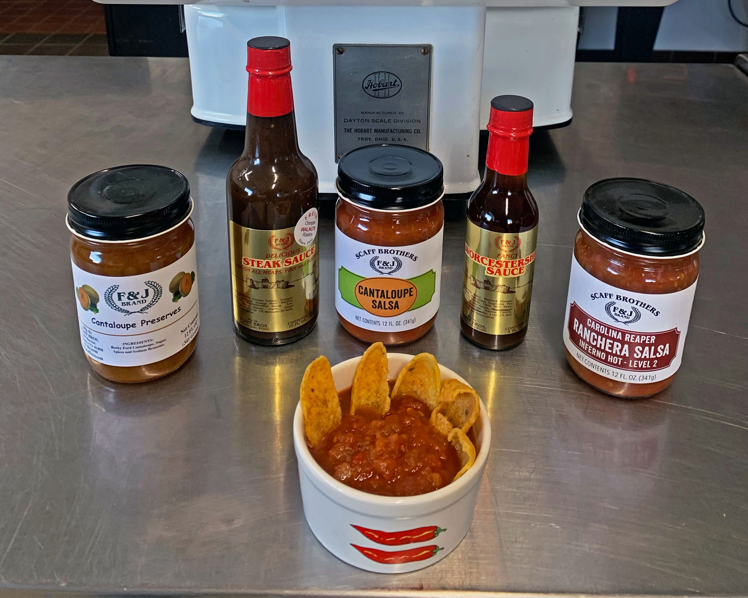 On a stainless steel table sits a bowl of salsa with frito-like chips in it. The bowl is surrounded by salsa, sauces, preserves from Scaff Brothers in La Junta.